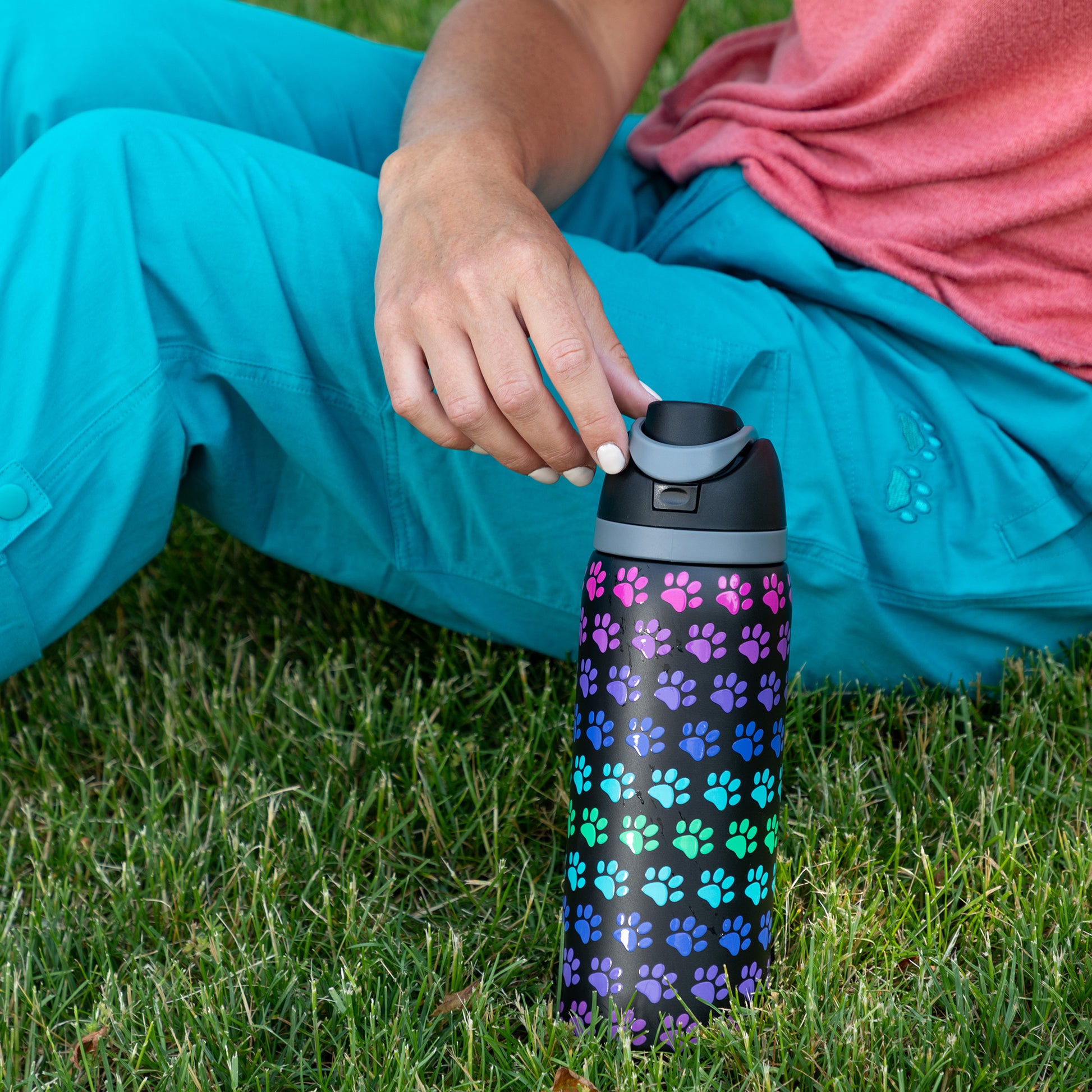 Person sitting on grass, holding a colorful, paw-patterned water bottle.