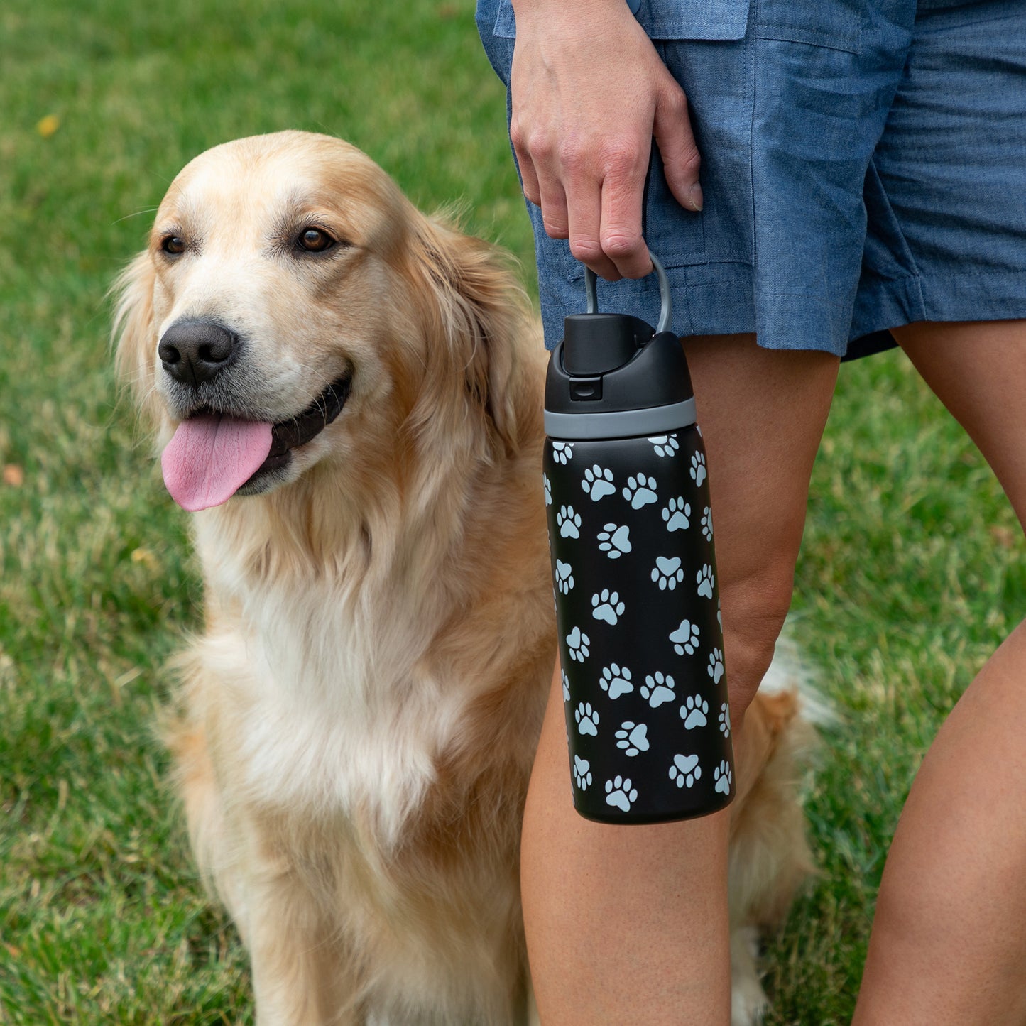 A golden retriever sits beside a person holding a paw-print water bottle.