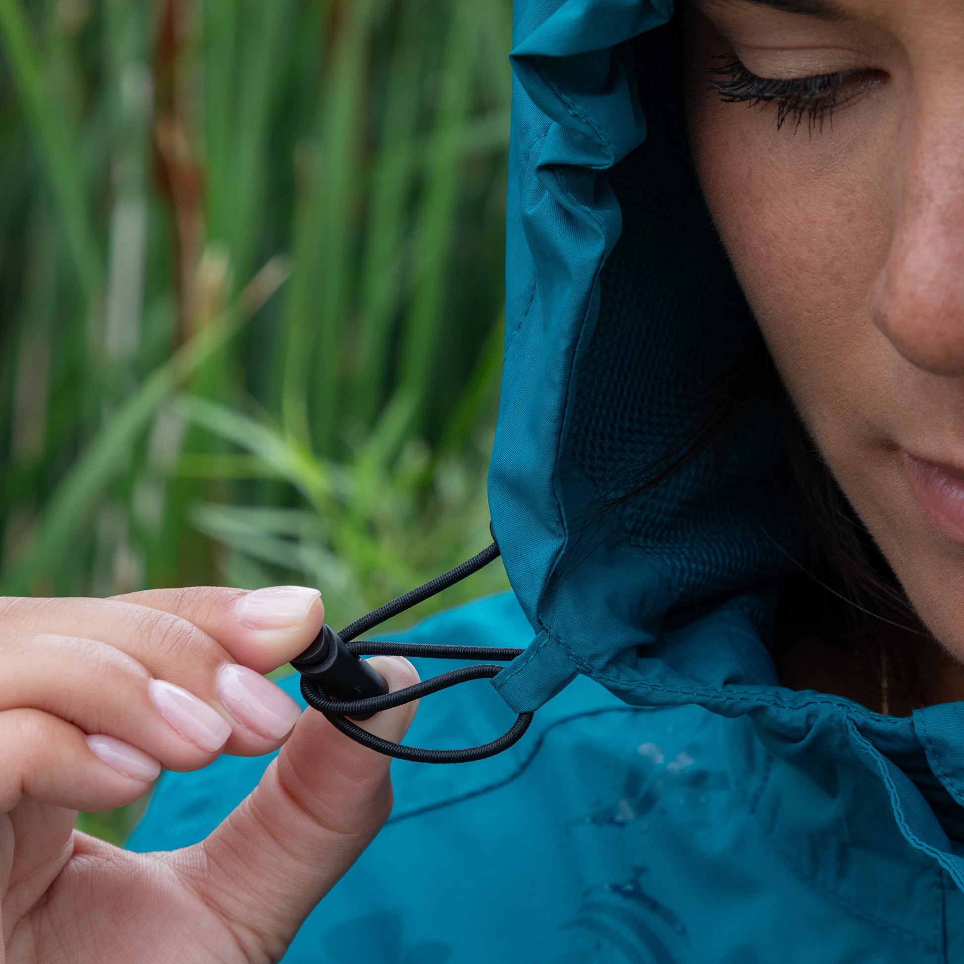 Person adjusting the hood of a blue raincoat with a blurred green background.