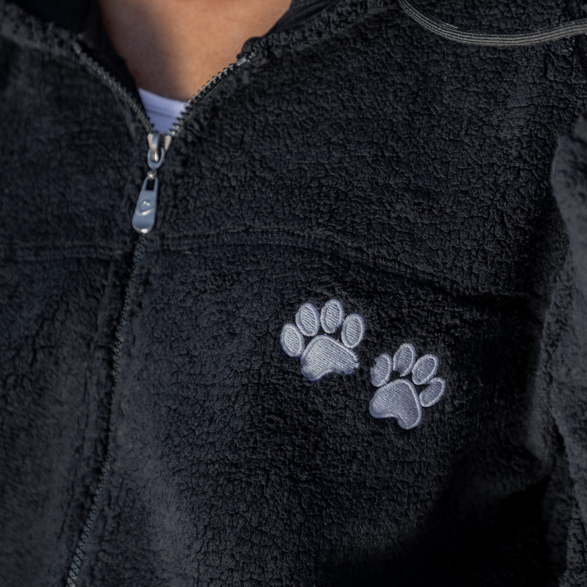 Close-up of a black fleece zip-up jacket with two embroidered gray paw prints on the chest, worn by a person.