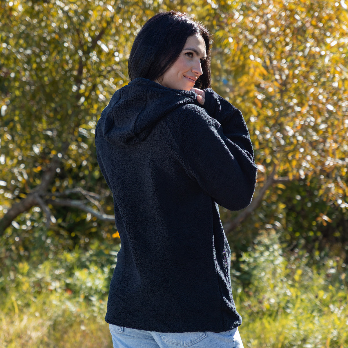 A woman wearing a black fleece hooded jacket, shown from the back and side, standing outdoors in front of a blurred background of green and yellow autumn trees.