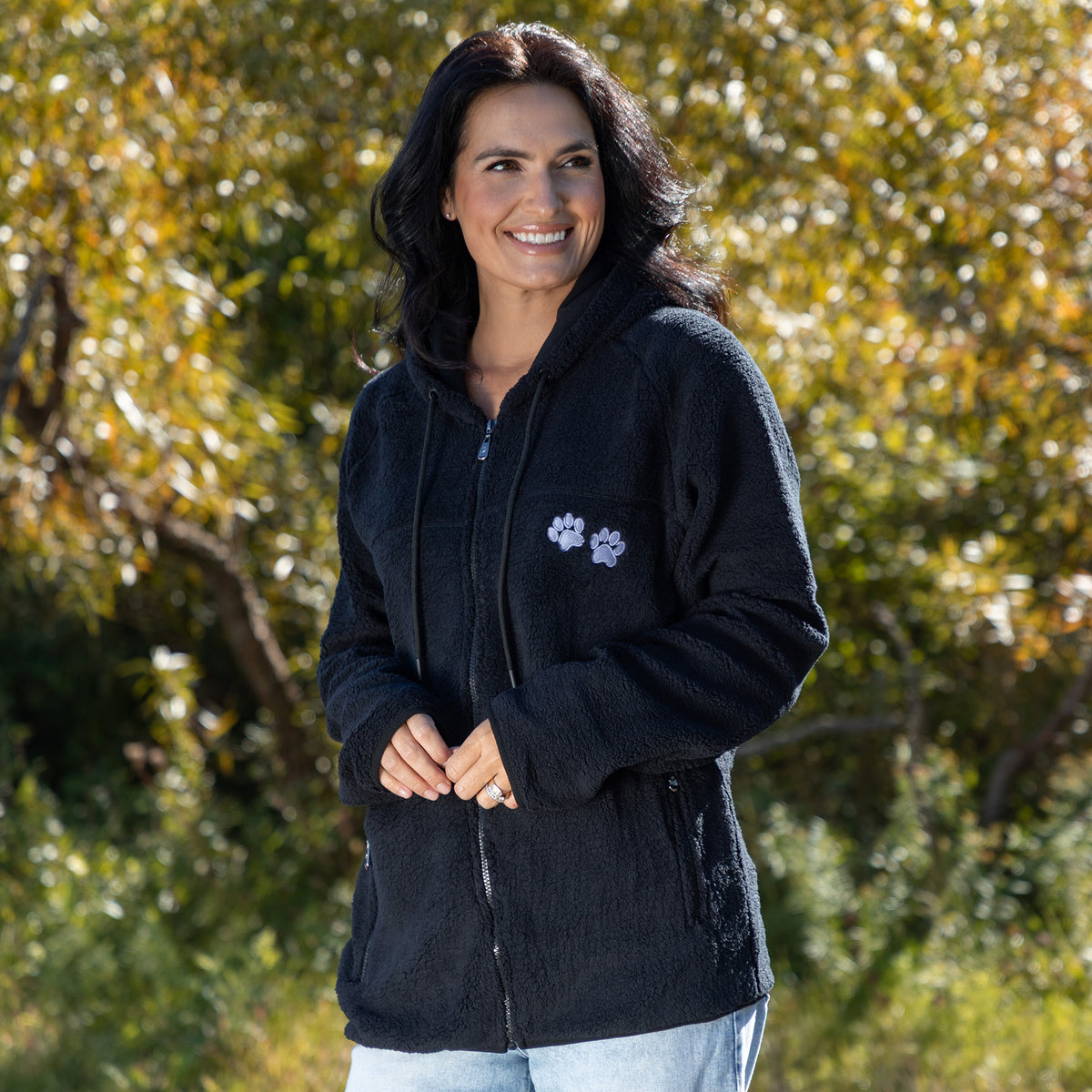 Smiling woman in a black fleece hoodie, surrounded by greenery and autumn foliage.