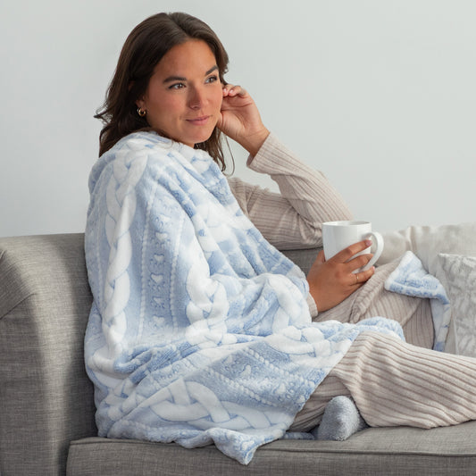 Woman sitting on a couch wearing a blue patterned blanket and holding a mug.