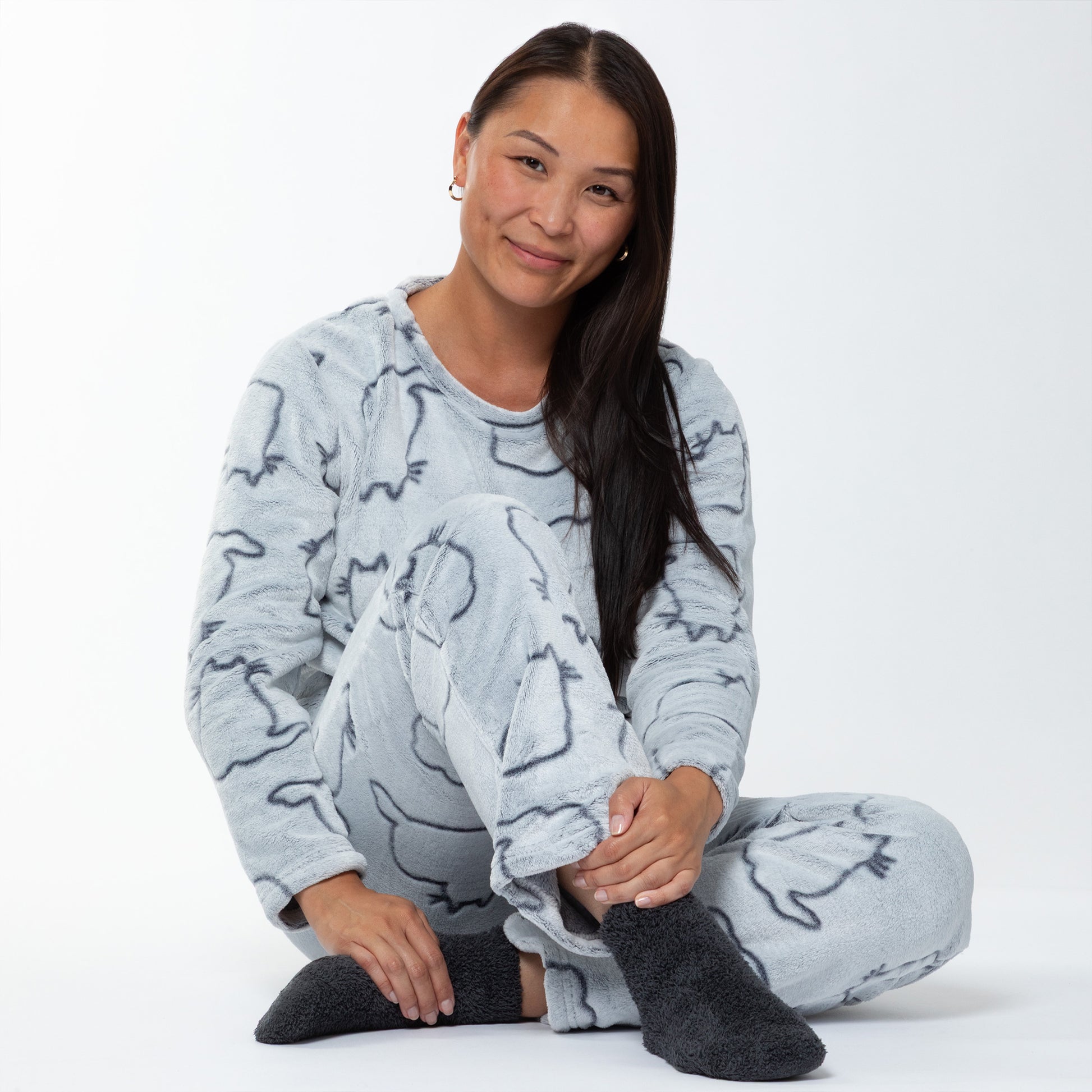A woman sitting in a relaxed position. She is wearing a matching set of fluffy, soft long-sleeve pajamas. The top and pants are light grey and patterned with cute cat outlines. 