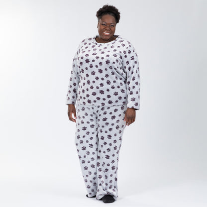 A woman wearing a matching set of fluffy, soft long-sleeve pajamas. The top and pants are light grey and patterned with purple paw prints. Plus and extended size available. 