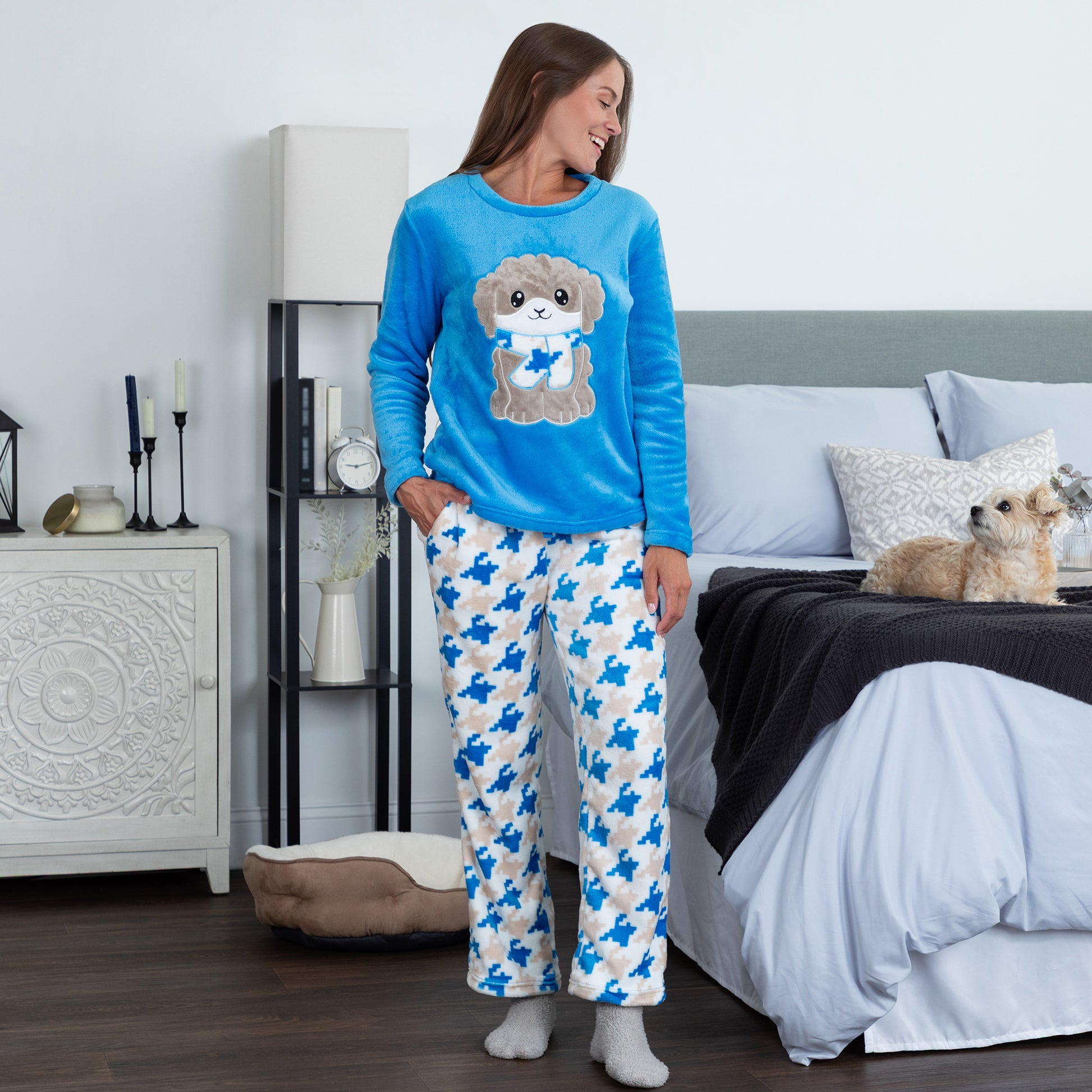 A woman wearing a blue long-sleeve top with an embroidered cute dog design and gray, blue and light blue houndstooth pattern. Design a blue dog in super cozy fabric, standing in a bedroom setting looking at a dog 