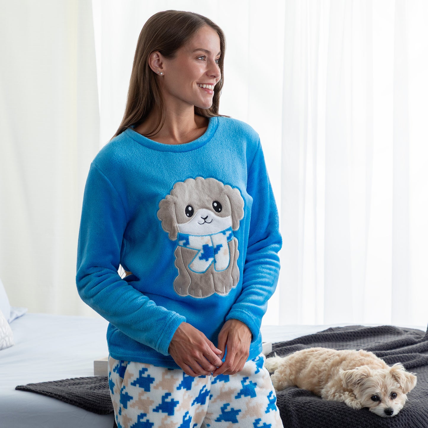 A woman in a cozy bedroom with her pet dog. She is wearing a blue long-sleeve top with an embroidered cute dog design and gray, blue and light blue houndstooth pattern. Design a grey dog with a scarf in super cozy fabric.