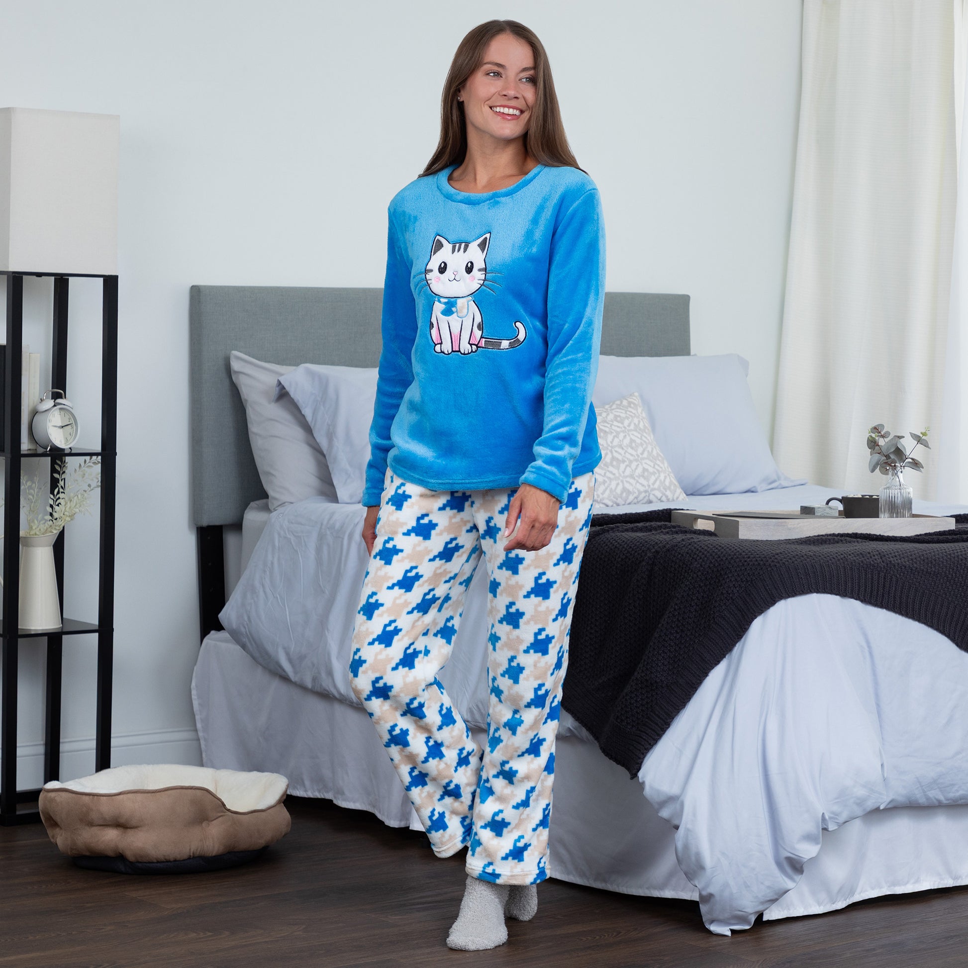 A woman wearing a blue long-sleeve top with an embroidered cute cat design and gray, blue and light blue houndstooth pattern. She is standing in a bedroom setting. Design a blue cat in super cozy fabric, showing an Elastic waistband with a drawstring, two side pockets