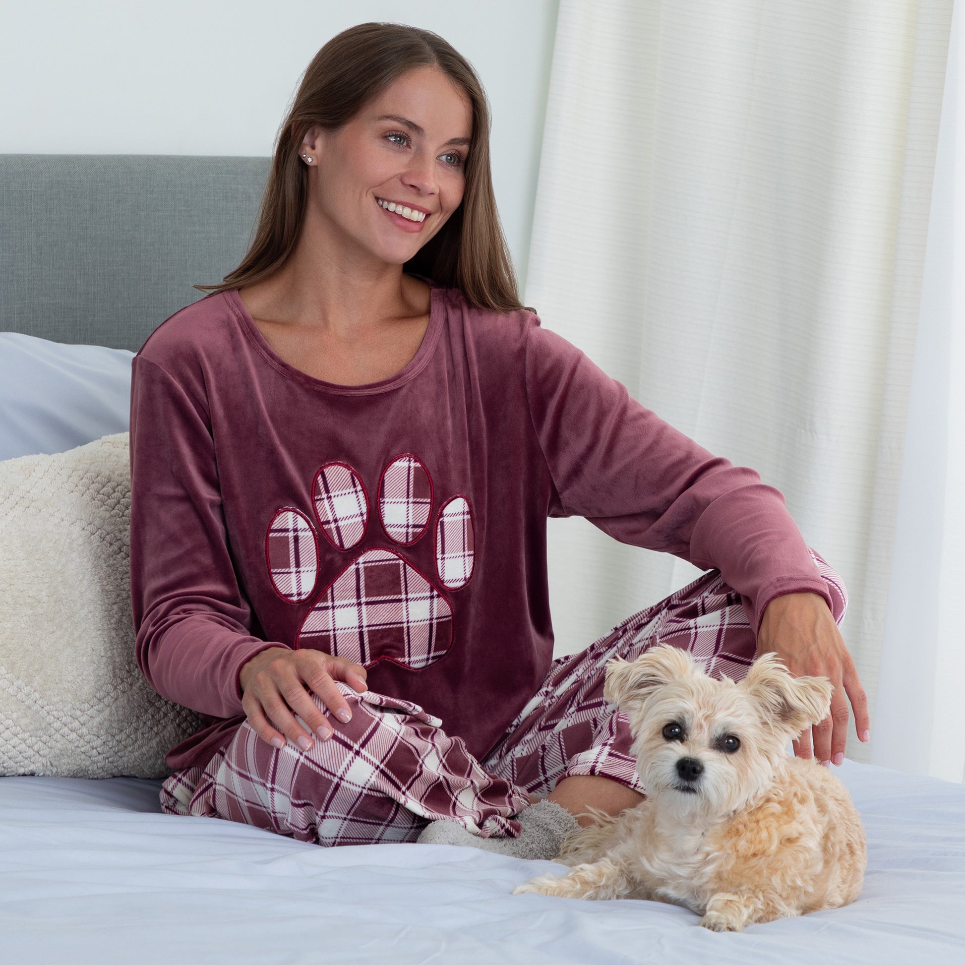Woman sitting in cozy bed with her pet dog. She is wearing a velour pajama set— the top is a maroon long-sleeve shirt with a large plaid paw print. The pants are the same white and maroon plaid pattern as the paw print on the shirt. 