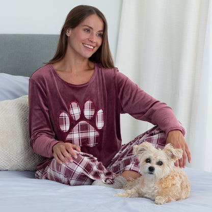 Woman sitting in cozy bed with her pet dog. She is wearing a velour pajama set— the top is a maroon long-sleeve shirt with a large plaid paw print. The pants are the same white and maroon plaid pattern as the paw print on the shirt. 