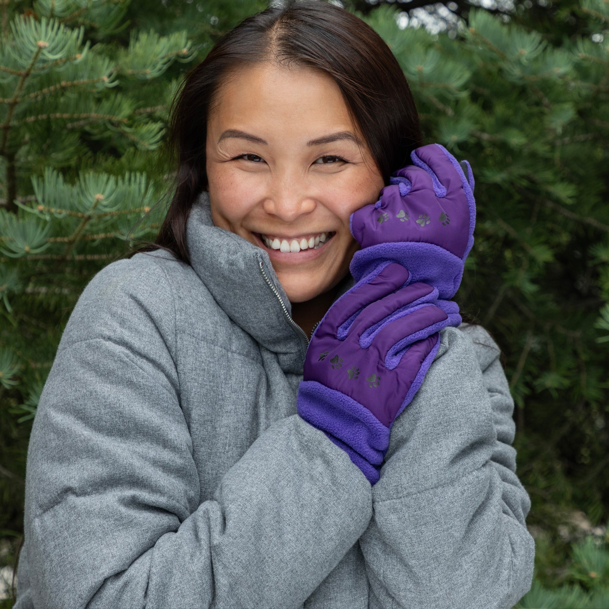 Woman wearing purple gloves and a gray coat in front of green trees