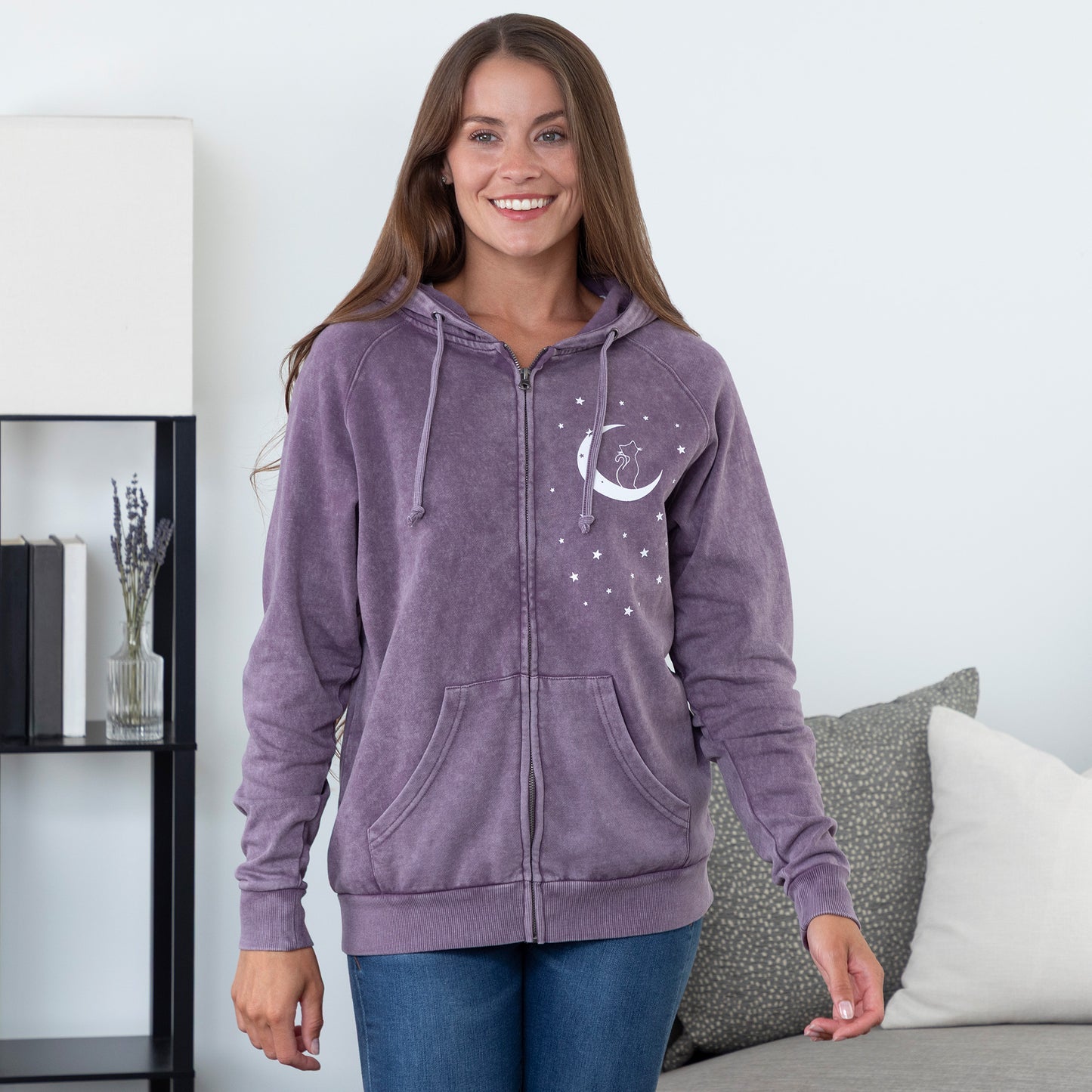Woman wearing a purple hoodie with a moon and star design, standing in a room with a white wall and a shelf.