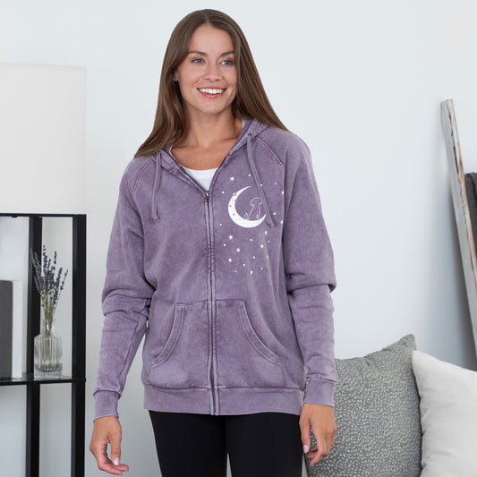 Woman wearing a purple hoodie with a moon and star design, standing in a room with a white wall and some decor items.