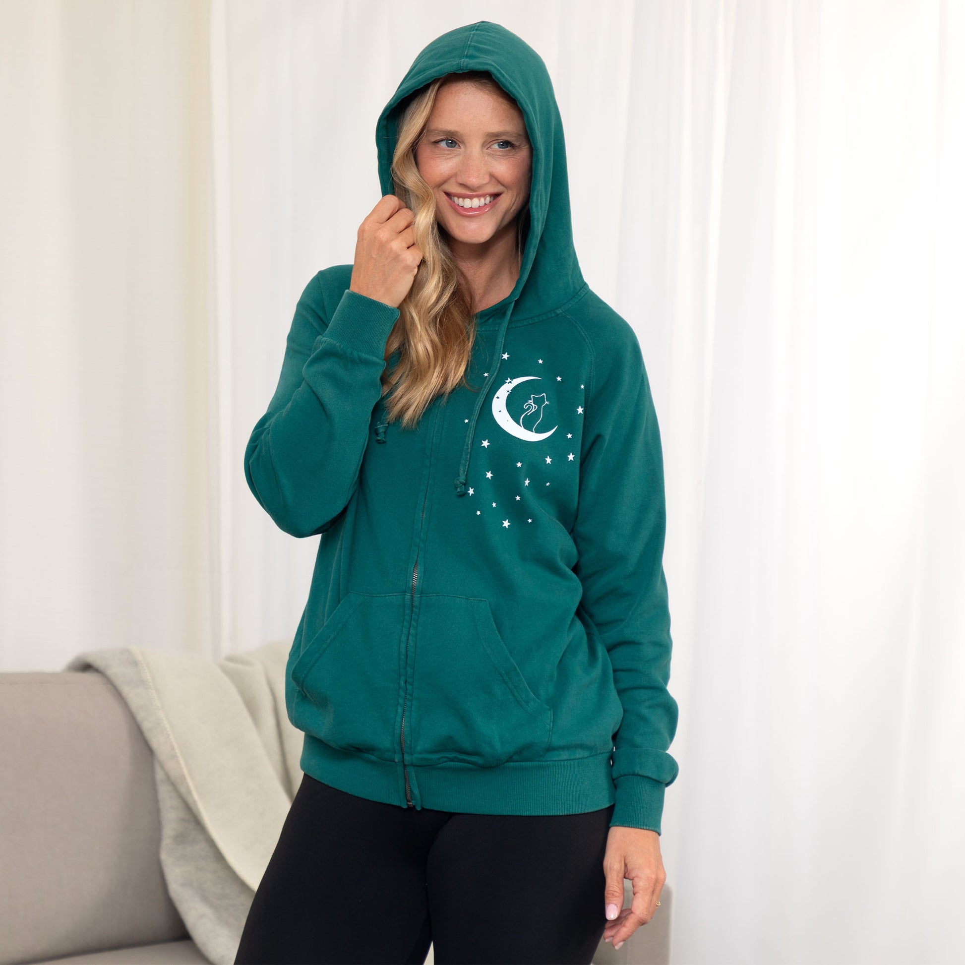 A woman wearing a teal green zip-up hoodie with a white crescent moon and stars graphic on the left chest, standing in front of white curtains.