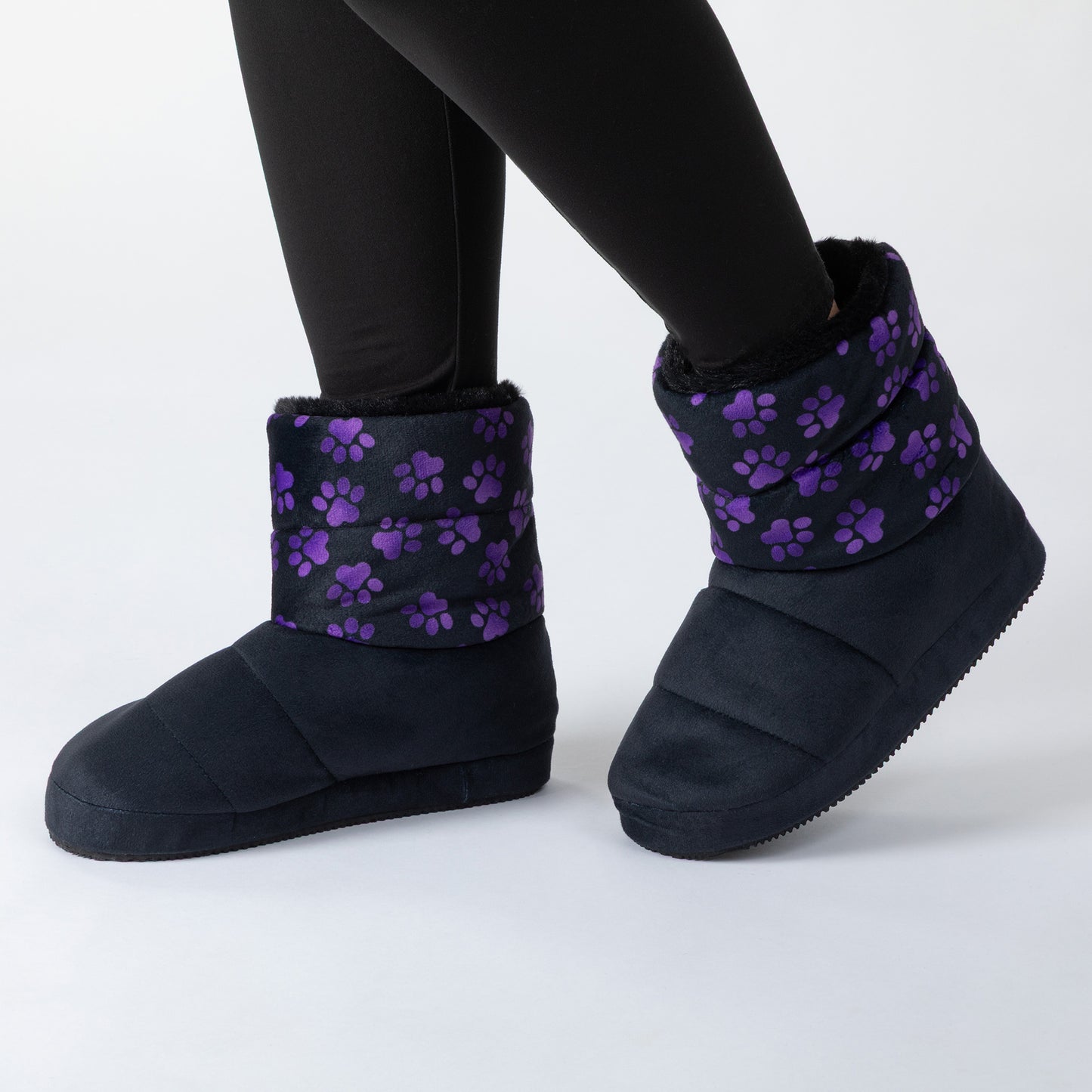 A person wearing black slip-on slipper boots with purple paw print patterns on the cuffs, standing on a white background.