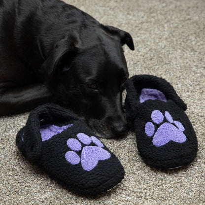 Black dog lying next to black slippers with purple paw prints on a carpeted floor.