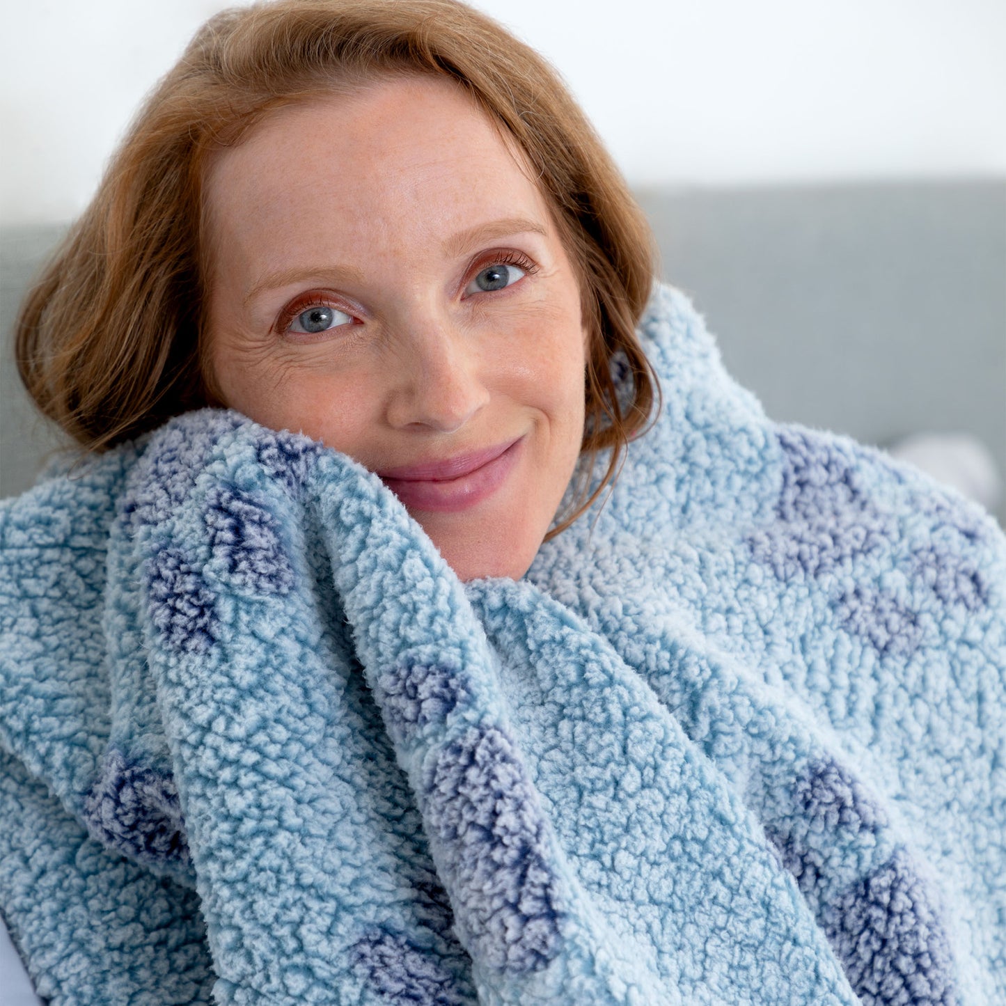 A close-up of a smiling woman wrapped in a soft, textured fleece blanket with a light blue and lavender pattern.