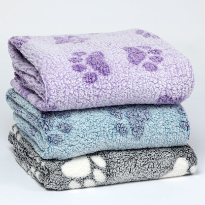 Three folded plush fleece blankets stacked on a white background, each with a purple paw print pattern. The top blanket is lavender, the middle is light blue, and the bottom is gray and white.