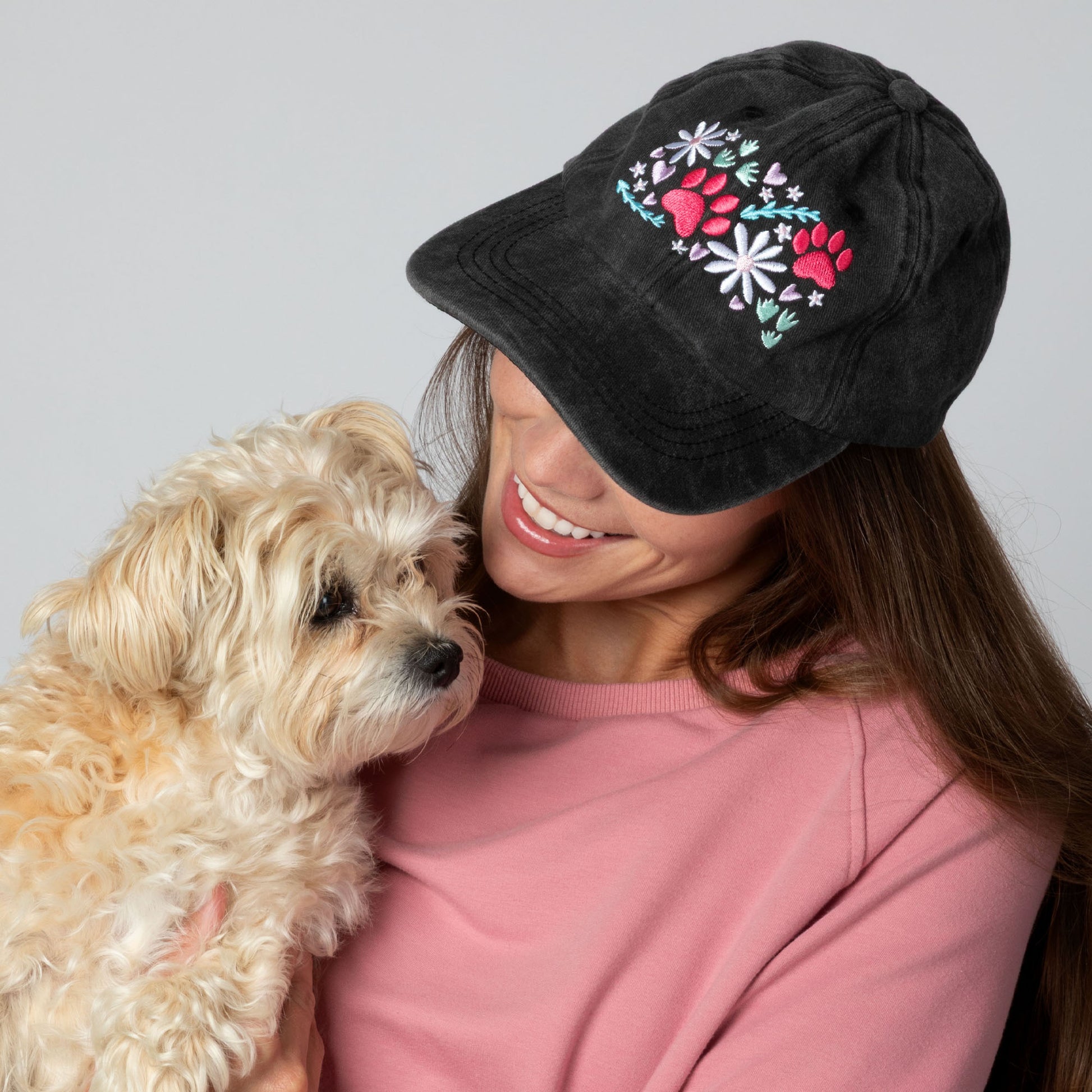Woman wearing a black cap with floral designs holding a dog against a gray background