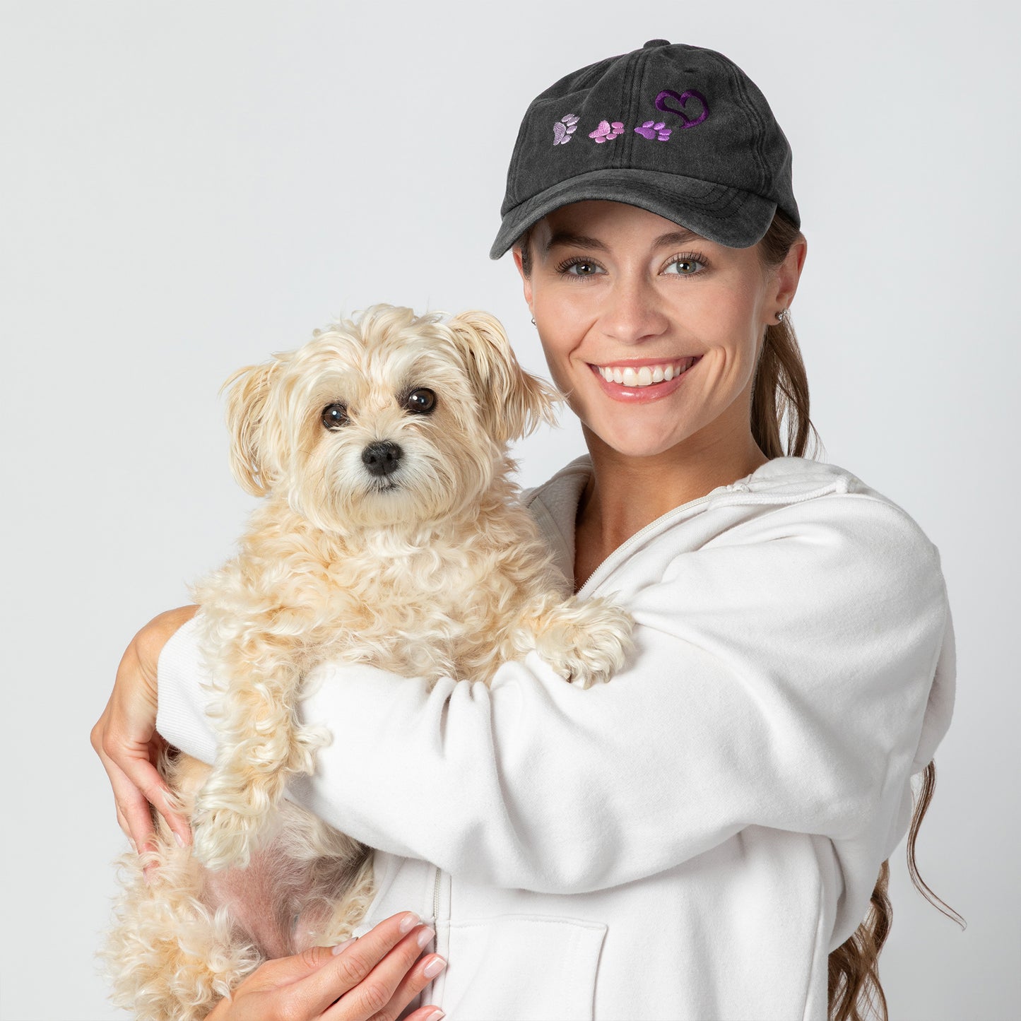 Woman holding a small white dog wearing a dark cap with paw prints and a heart on a light gray background