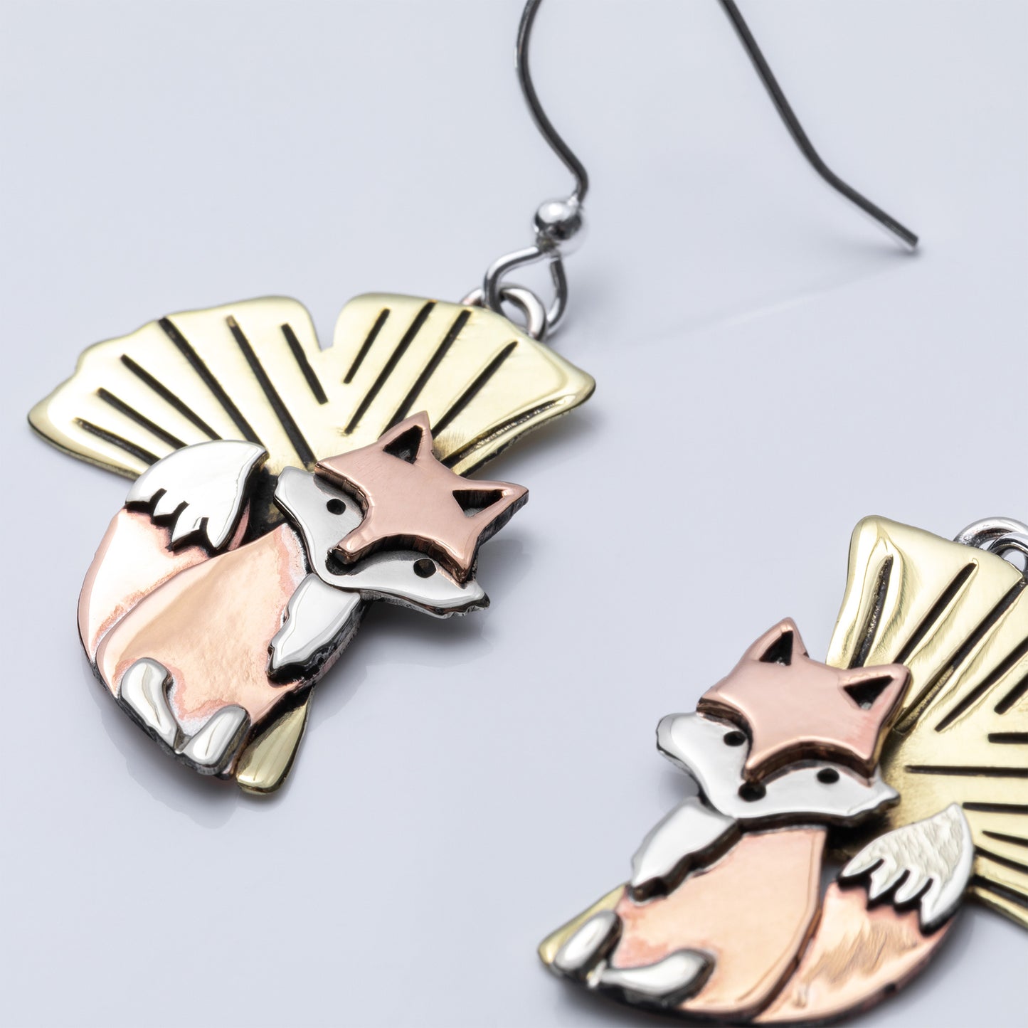 Pair of earrings with fox and leaf design in copper, silver, and brass, on a light gray background.