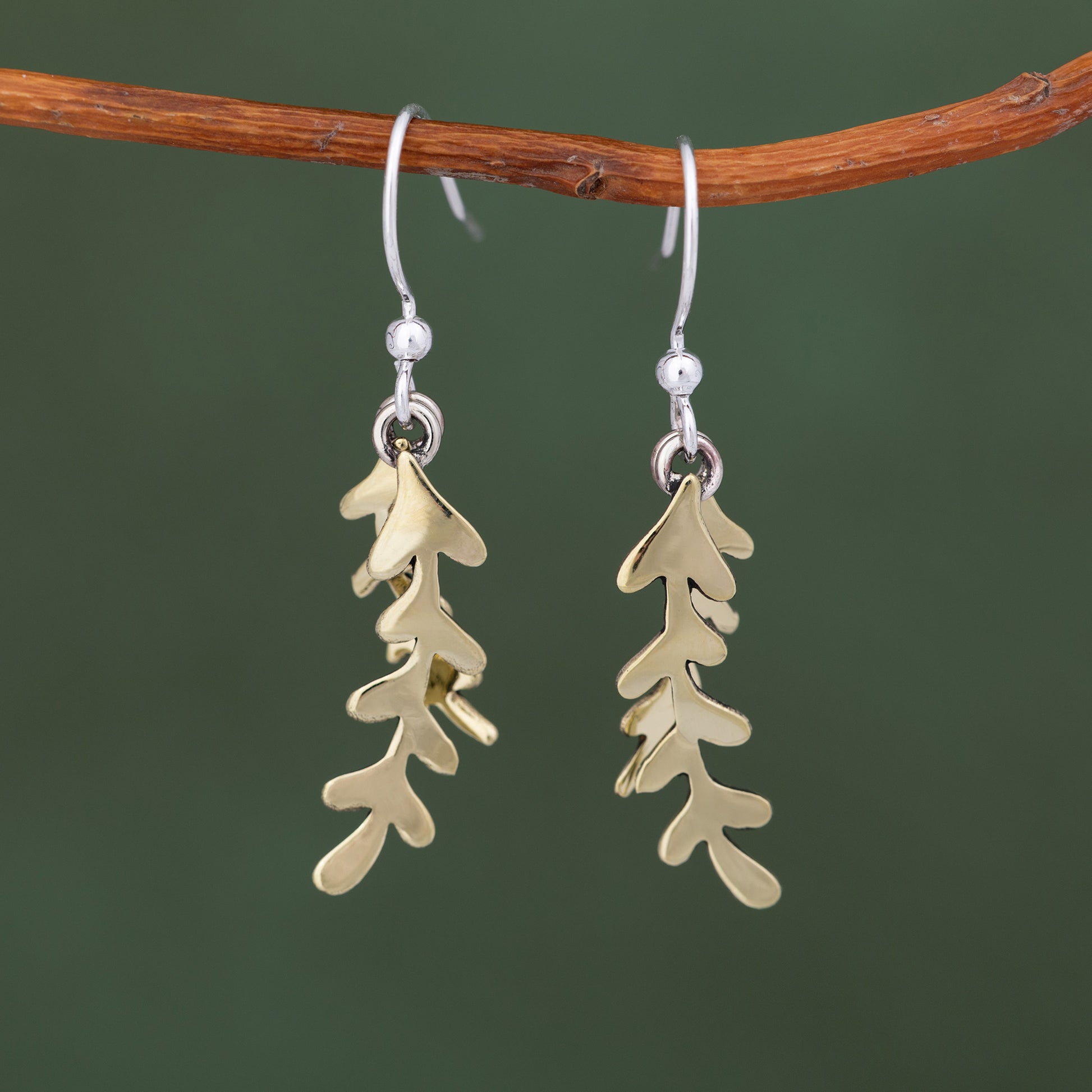 Brass leaf-shaped earrings hanging from a branch against a green background