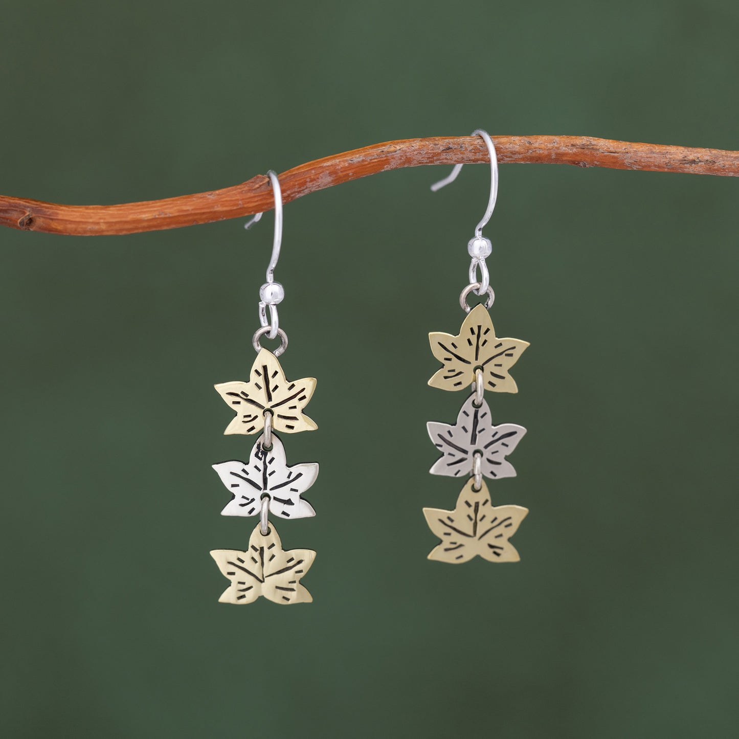 Pair of leaf-shaped earrings in silver and brass on a branch against a green background