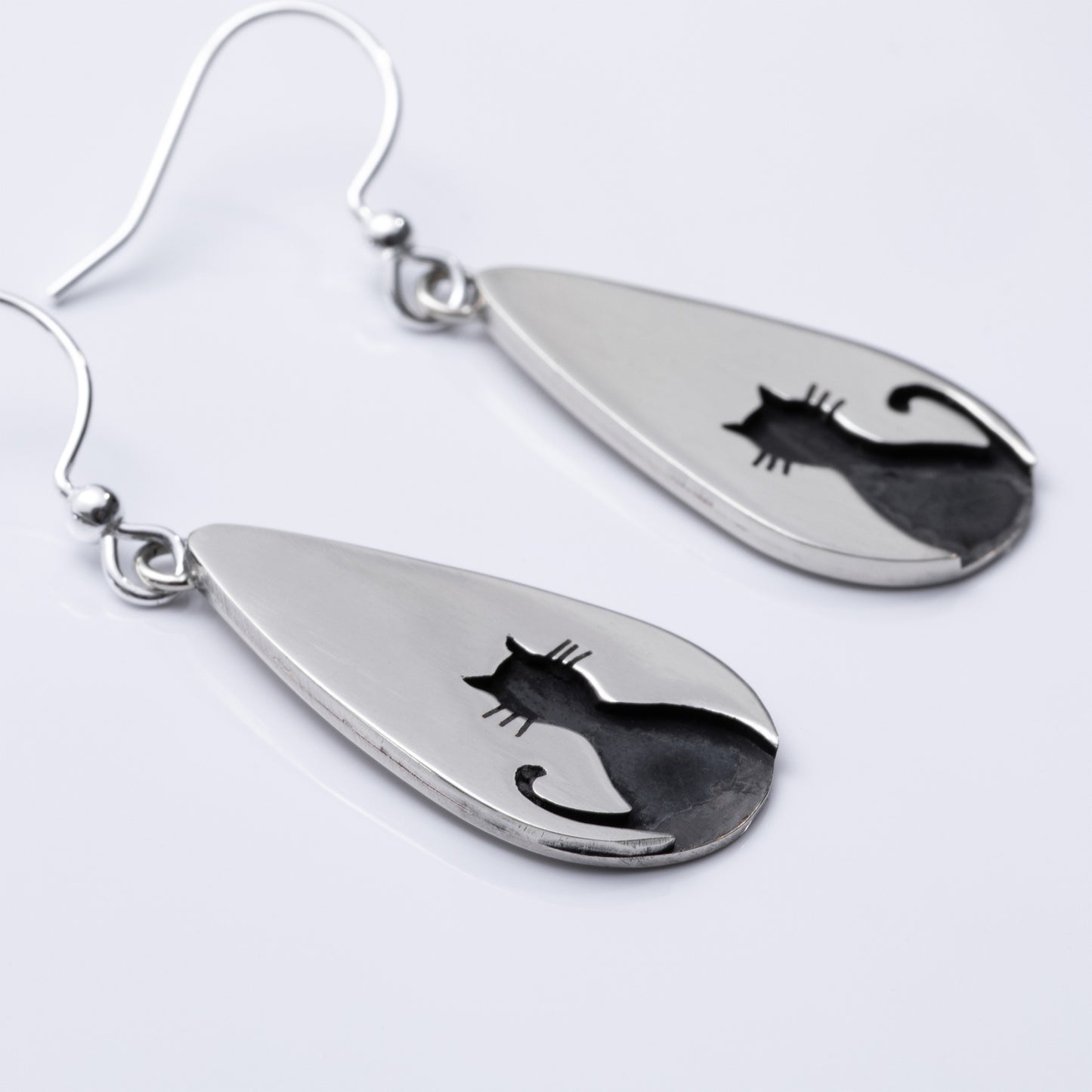 Close up of teardrop-shaped silver earrings with cat silhouettes on a light gray background