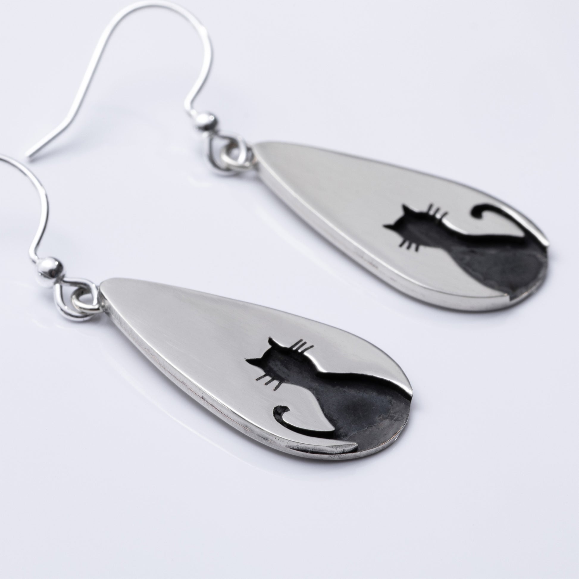 Close up of teardrop-shaped silver earrings with cat silhouettes on a light gray background