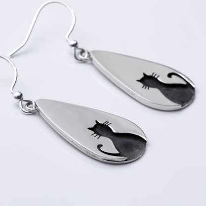 Close up of teardrop-shaped silver earrings with cat silhouettes on a light gray background