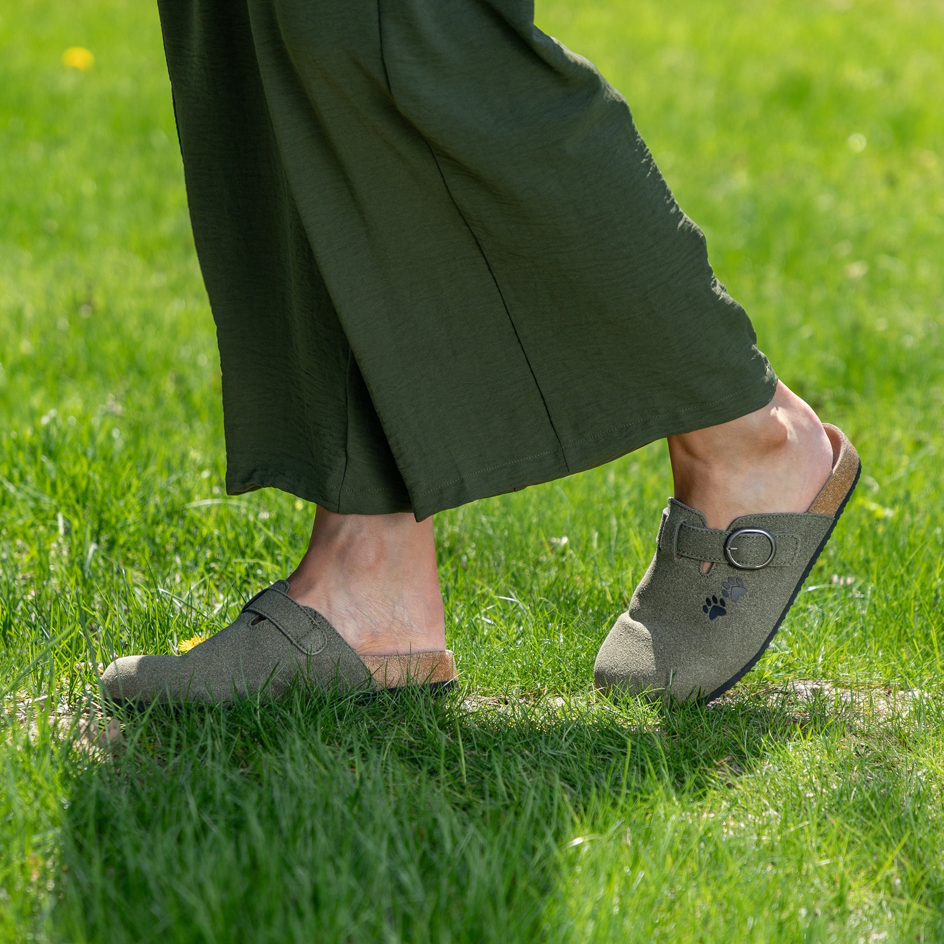 Woman walking in grassy area, wearing green clogs and loose olive pants.