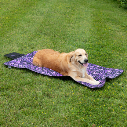 A golden retriever dog lies on a purple patterned outdoor folding blanket on a green grass lawn.