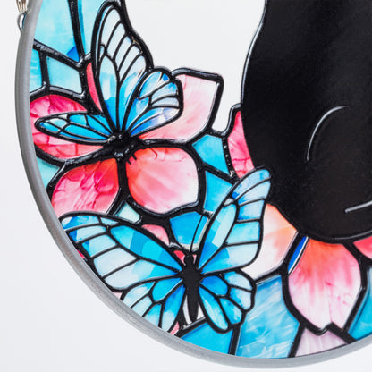 Close-up of a sun catcher in a stained-glass-style butterfly and flower design in blue, pink, and light blue with black outlines, alongside a black curved accent on a white background.
