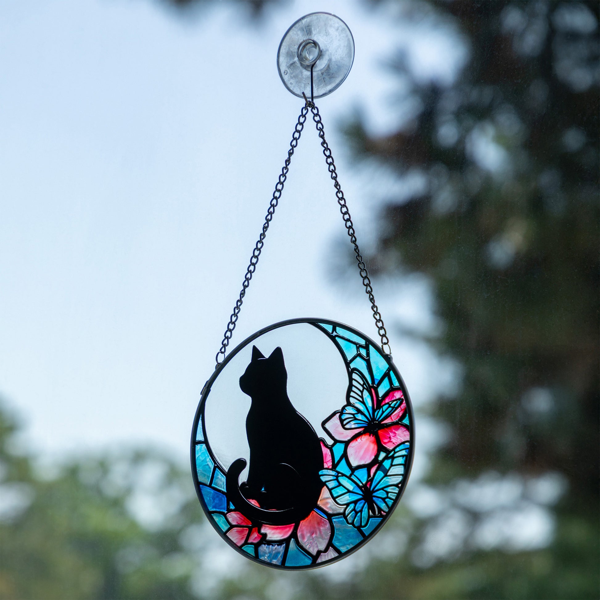 An oval stained-glass-style hanging ornament with a black cat silhouette, pink flowers, and blue butterflies, suspended by chains from a clear suction cup against an outdoor blurred background.