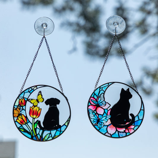 Two circular stained-glass style hanging suncatchers with black cat and dog silhouettes, displayed outdoors against a blurred sky and tree background. One features a black dog with yellow and orange flowers and a butterfly, and the other features a black cat with pink and blue flowers and a butterfly.