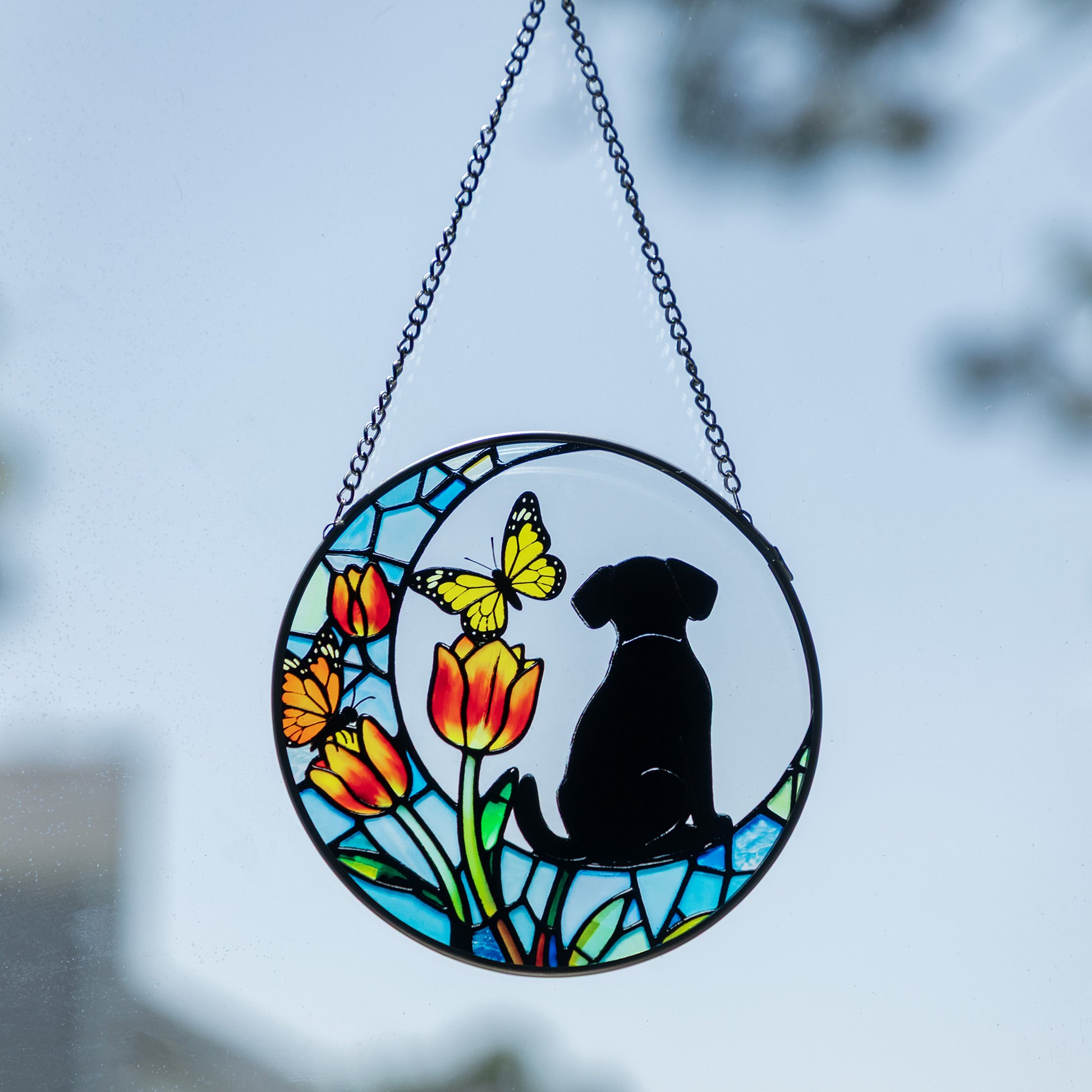 A circular stained-glass-style hanging ornament with a black dog silhouette, yellow and orange tulips, and a butterfly, suspended by a chain against a blurred outdoor sky background.