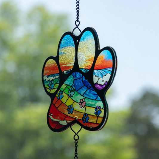 A stained glass paws print suncatcher hanging from a black metal chain against a blurred green outdoor background.
