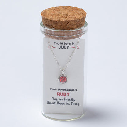 Necklace with a red gemstone in a glass jar with a cork lid, featuring text about July-born individuals and their birthstone.
