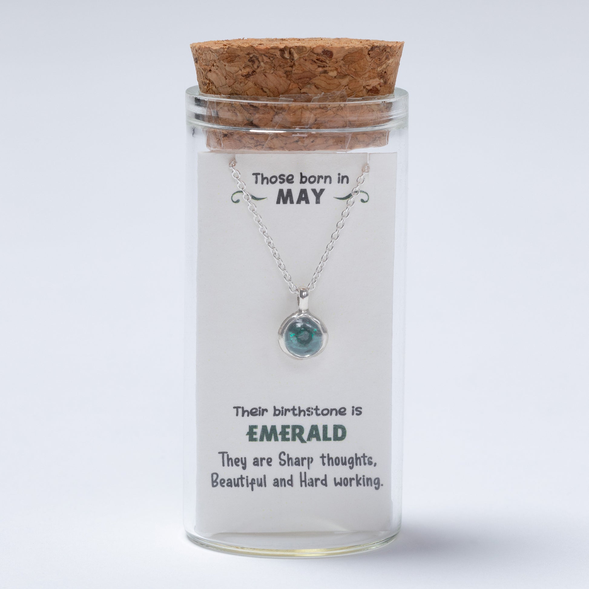 Necklace with a pendant in a glass jar labeled for May birthstone, on a light gray background
