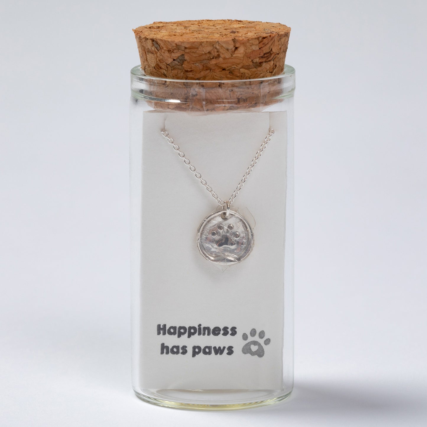 Glass jar with cork lid containing a necklace with a paw print pendant, on a light gray background.