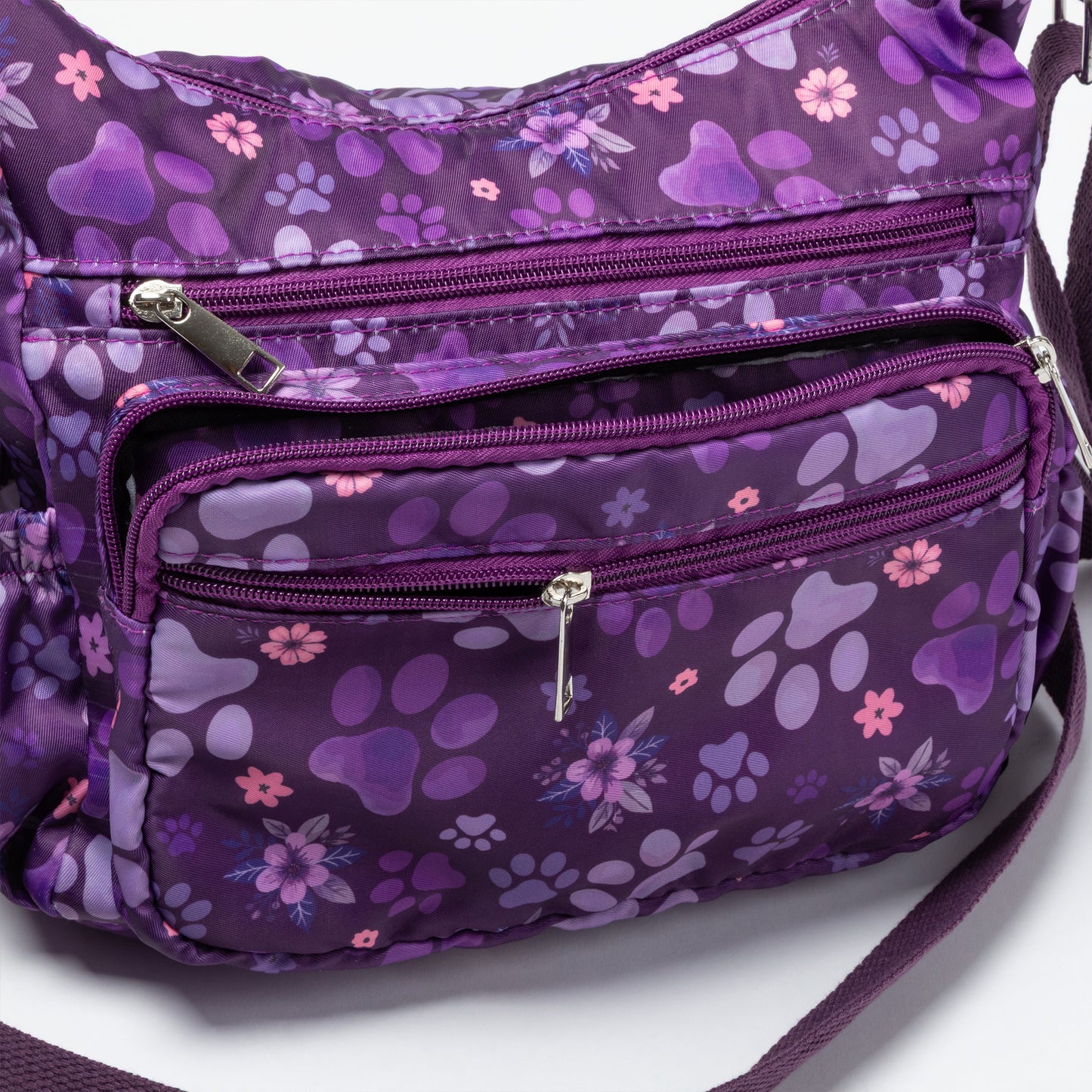 A purple nylon crossbody bag with a floral and paw print pattern, featuring multiple zippered compartments and a silver zipper pull.