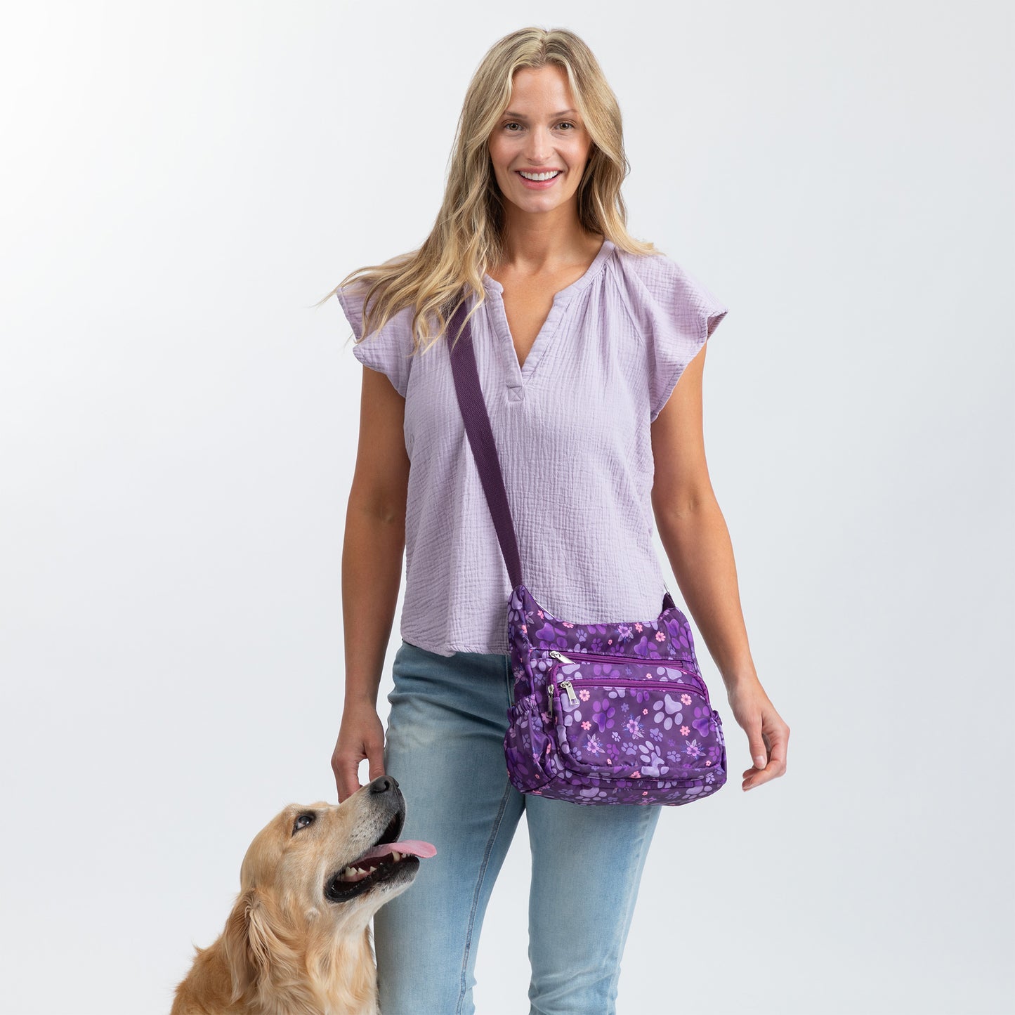 A woman wearing a light purple blouse and blue jeans carries a purple paw crossbody bag with a matching strap. A golden retriever dog is standing on the left side of the image against a white studio background.