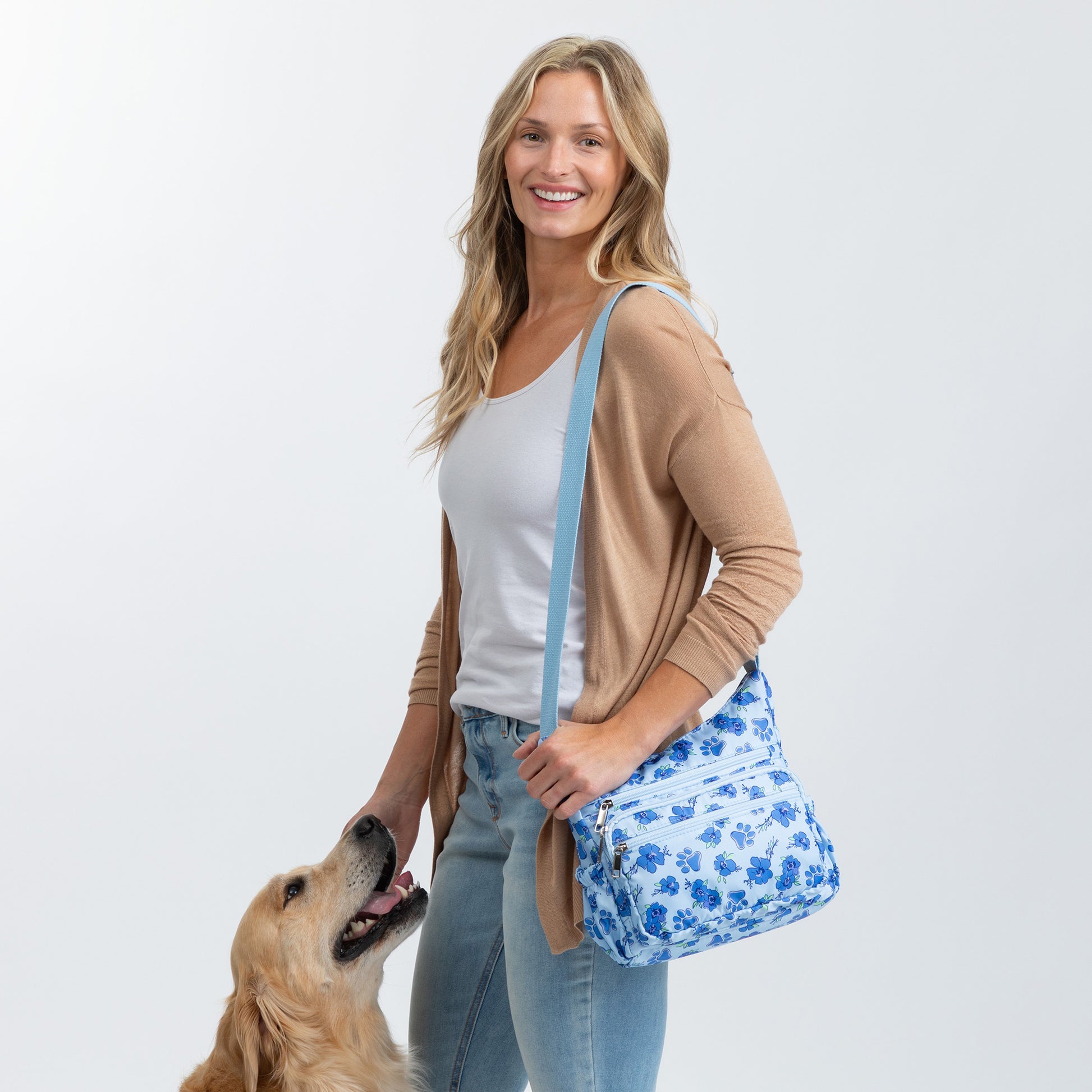 A woman wearing a tan cardigan and blue jeans carries a blue floral crossbody bag with a light blue strap, standing next to a golden retriever dog against a light gray background.