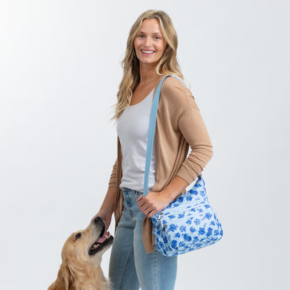 A woman wearing a tan cardigan and blue jeans carries a blue floral crossbody bag with a light blue strap, standing next to a golden retriever dog against a light gray background.