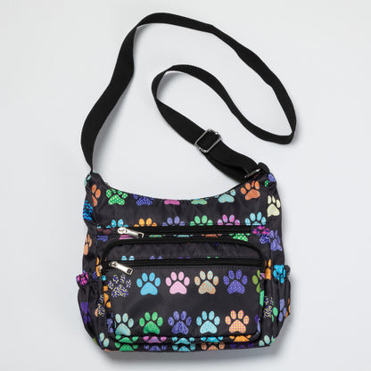 A black nylon crossbody bag featuring a colorful pattern of dog paw prints in various colors, with a black adjustable shoulder strap and silver zipper hardware.