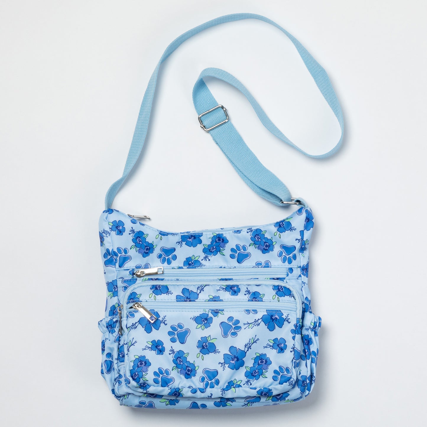 A light blue crossbody bag with a medium blue floral pattern, featuring multiple zippered pockets and an adjustable shoulder strap, shown against a plain white background.