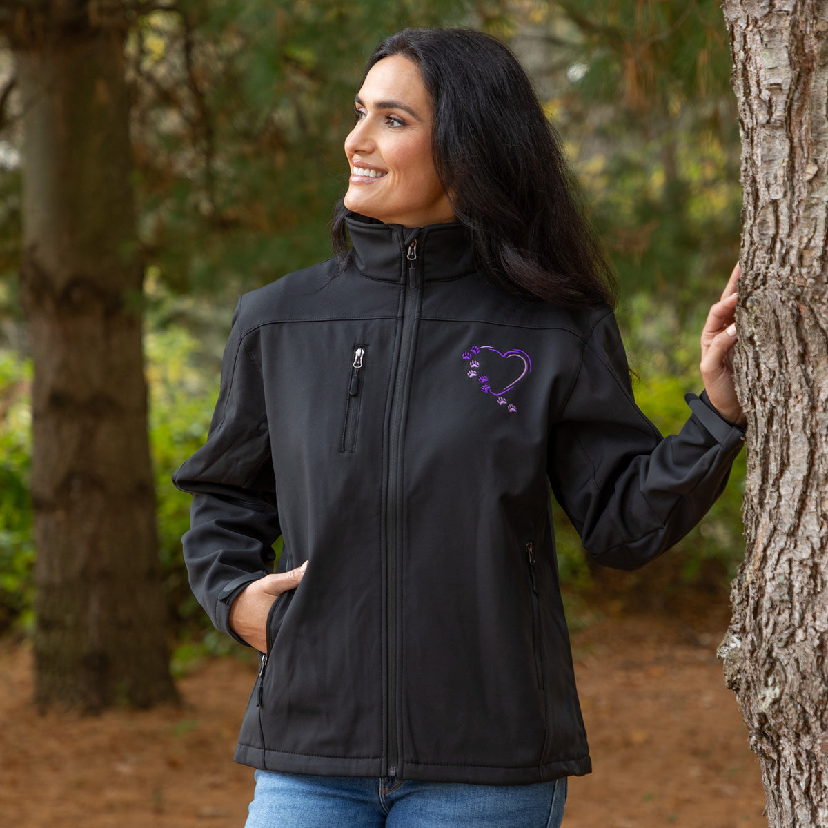 A woman wearing a black softshell jacket with a purple heart-shaped arrow graphic on the chest, standing outdoors next to a tree.