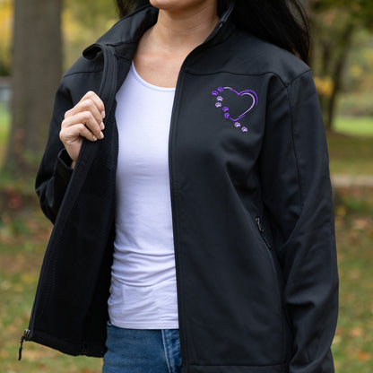 A woman wearing a black zip-up jacket outdoors, with a purple heart graphic and small paw prints on the left chest, layered over a white tank top and blue jeans.