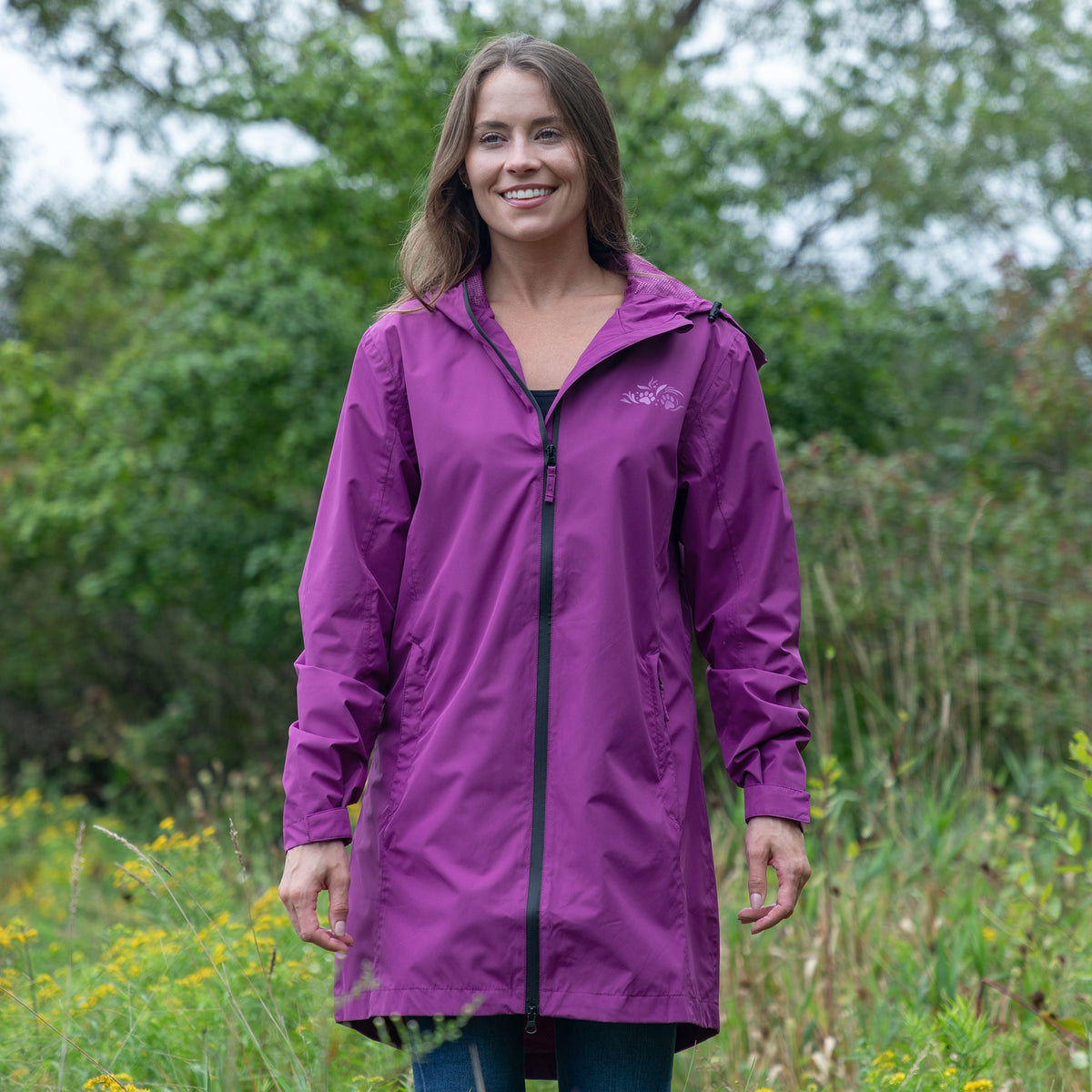 A woman wearing a purple hooded raincoat with a black front zipper, standing outdoors in a grassy field with trees in the background.