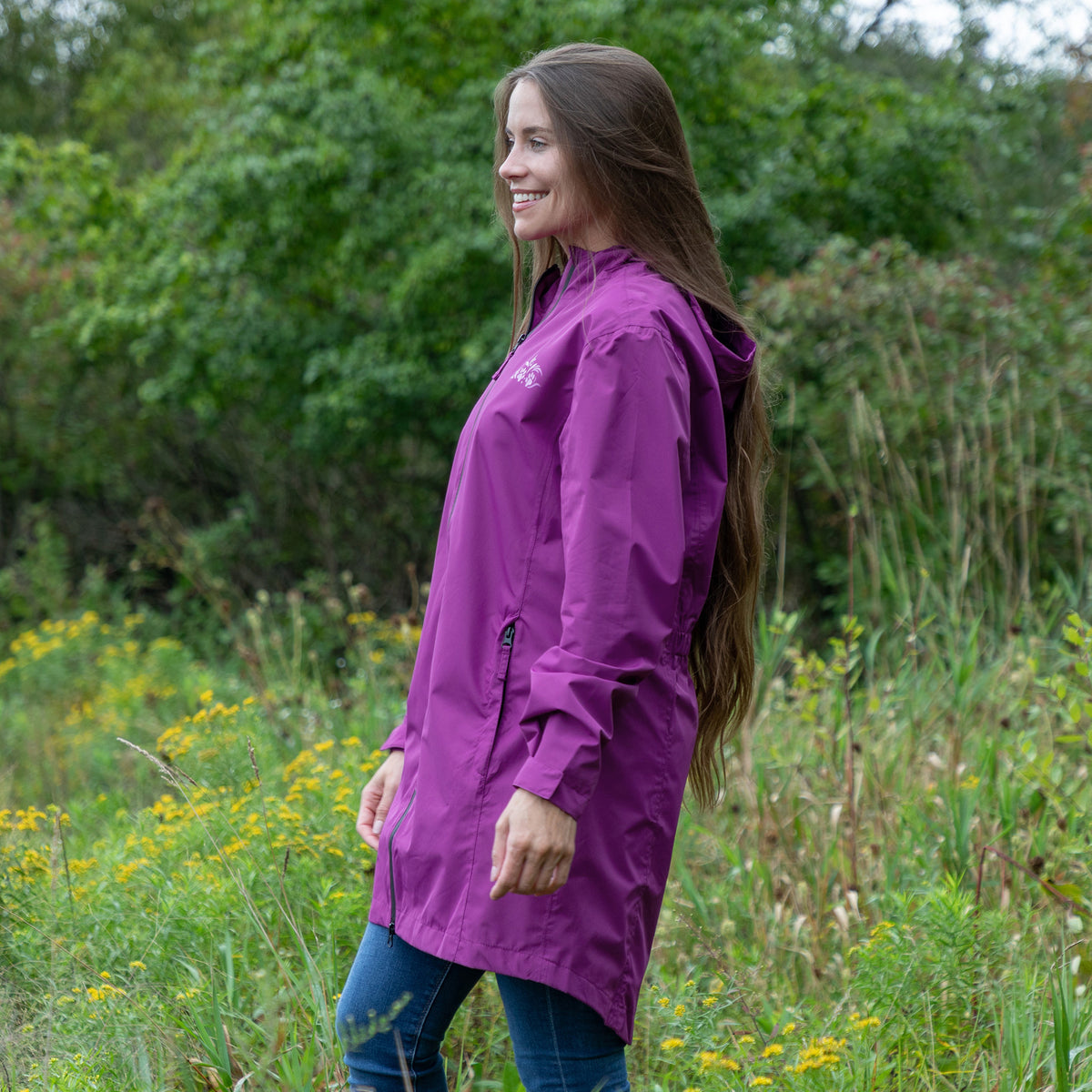 A woman wearing a purple hooded raincoat and blue jeans standing outdoors in a grassy field with green trees in the background.
